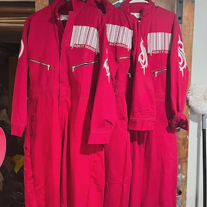 Könnte beinhalten: Drei leuchtend rote Overalls, die an weißen Kunststoffbügeln hängen. Jeder Overall hat eine weiße Strichcode-Grafik auf der Brust mit dem Text 42617 00002, ein weißes Tribal-Logo auf dem Ärmel und horizontale Reißverschlusstaschen auf der Brust.