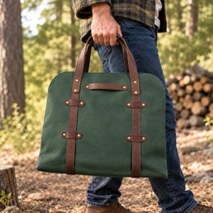 Op de afbeelding: Een bosgroene canvas tas met twee verticale bruine leren riemen met koperen klinknagels, twee handvatten aan de bovenkant en een horizontaal bruin leren handvat in het midden van het voorpaneel. De tas heeft een rechthoekige vorm met afgeronde hoeken.