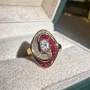 Puede incluir: Un anillo de oro con un diamante central de talla oval rodeado por un diseño en espiral de rubíes y diamantes talla baguette engastados en canal. Las piedras están dispuestas en un patrón curvo y superpuesto en una banda de oro amarillo pulido.