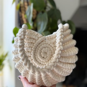 May include: Cream-colored, hand-knitted basket with a spiral design. The basket features a central circular pattern and a textured, ridged exterior. The rim is finished with a braided rope detail. A decorative and functional item.