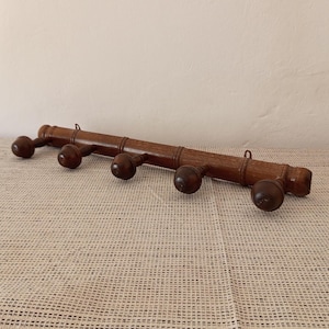 May include: A horizontal wooden wall rack featuring five rounded pegs for hanging items. The rack is crafted from medium-toned brown wood with a smooth, polished finish and includes two small metal loops on the top edge for wall mounting.