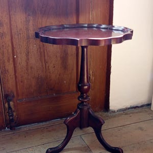 Puede incluir: Una mesa de pedestal de madera de color marrón oscuro con una superficie redonda de borde festoneado. La mesa se apoya en una columna central torneada con tres patas curvas que se extienden hacia afuera formando una base de trípode estable.