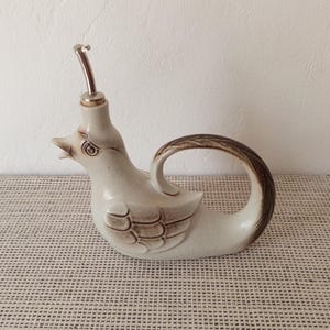 Puede incluir: Una aceitera de cerámica con forma de pato, acabado en color crema moteado y detalles en marrón en las alas y la cola. El pato presenta detalles moldeados en el pico y los ojos, con un vertedor metálico de color plateado en la abertura.