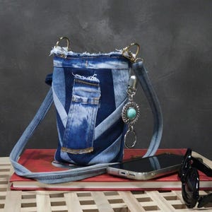 Peut inclure: Un sac bandoulière fabriqué à partir de tissu denim bleu recyclé avec un bord supérieur effiloché et une poche verticale. Le sac comprend une longue bandoulière en jean et un accessoire à clip en métal argenté avec une pierre turquoise.