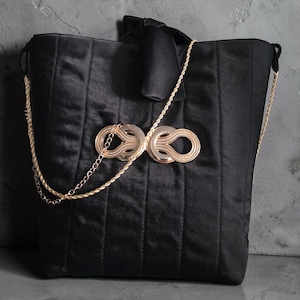 Peut inclure: Un sac cabas noir matelassé avec des coutures verticales, un nœud sur le dessus et une bandoulière en chaîne dorée. L'avant est orné d'un fermoir décoratif en métal doré composé de deux boucles circulaires entrelacées.