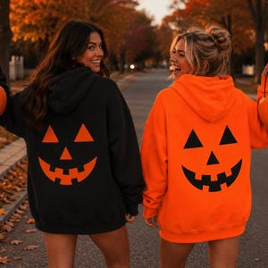 Puede incluir: Una sudadera con capucha negra con un estampado grande de cara de calabaza naranja en la espalda y una sudadera con capucha naranja con un estampado grande de cara de calabaza negra en la espalda, cada una con un cubo de calabaza de plástico.