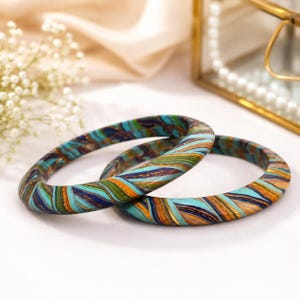 May include: Two circular bangle bracelets featuring a marbled pattern of turquoise, forest green, navy blue, and metallic gold swirls. The bracelets have a smooth, polished finish and a rounded profile, showcasing a continuous blend of colors.