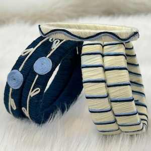 May include: A set of four circular bangles wrapped in thread. Two dark navy blue bangles feature light blue circular accents and white embroidered vine patterns. Two cream-colored bangles feature horizontal stripes in navy blue and light blue.