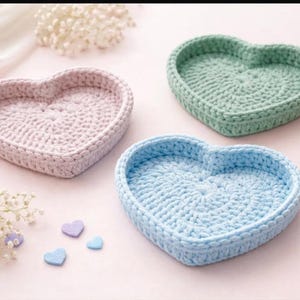 May include: Three handmade crochet heart-shaped trays in pastel colors: pink, green, and blue. The trays are arranged on a light pink surface with small heart-shaped embellishments. The crochet work is visible, showcasing the texture.