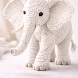 Peut inclure: Un éléphant en crochet de couleur crème doté de grandes oreilles texturées, d'une trompe, de deux petites défenses couleur ivoire et d'yeux en perles noires. La peluche présente un motif de mailles visible et une petite queue à pompon.