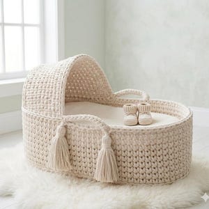 Può includere: Una culla per neonati all'uncinetto color crema, di forma arrotondata e con cappuccio. La culla presenta due manici e nappe decorative. Due piccoli stivaletti per neonati color crema sono all'interno della culla. La culla è posizionata su un tappeto bianco e soffice.