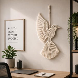 May include: A macrame dove wall hanging with outstretched wings, crafted from off-white cotton rope. The bird is suspended from a loop, and hangs against a neutral wall. A framed print with the words "FOCUS PLAN EXECUTE SUCCEED" is visible.