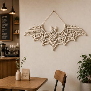 Könnte beinhalten: Ein cremefarbenes Makramee-Fledermaus-Wandbehang. Die Fledermaus hat ein detailliertes, symmetrisches Design mit strukturiertem Aussehen. Sie ist an einem Holzdübel an einer neutralfarbenen Wand aufgehängt.