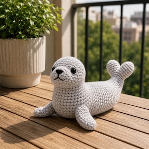May include: A crocheted seal toy in light grey, featuring black eyes and a smiling mouth. The seal is positioned on a wooden surface, with its flippers outstretched. The tail is heart-shaped. The toy is approximately 25 cm long.