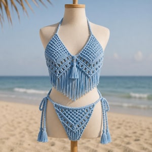May include: A light blue, handmade crochet bikini set. The halter top features a V-neck design with fringe and a tassel. The bikini bottoms have tie sides and tassels. The set is displayed on a mannequin against a beach backdrop.