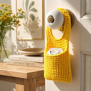 May include: A vibrant yellow, crocheted door tidy, containing white wireless earbuds. The tidy hangs from a white doorknob with brass fittings. The background features a wooden table, books, and a framed print with the word "MATISSE".