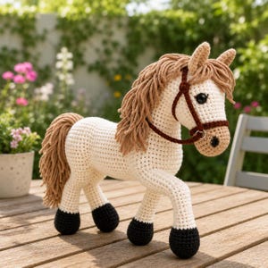 May include: A crocheted horse figurine in cream with a brown mane, tail, and bridle. The horse has black hooves and is posed on a wooden surface. The background features greenery and flowers, creating a natural setting for the handmade toy.