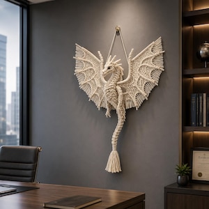 May include: A cream-coloured macrame dragon wall hanging with outstretched wings. The dragon has a detailed, textured design, with a long tail ending in a tassel. It is suspended from a wall by a cream-coloured cord.