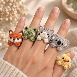 Puede incluir: Un conjunto de cinco anillos de ganchillo con diseños de animales que incluyen un zorro, una rana verde, un gato atigrado gris, un koala y un pollito amarillo. Cada anillo está hecho de hilo y muestra pequeños rasgos faciales y patas.