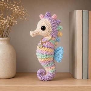 May include: A handcrafted crochet seahorse in pastel colors, including lavender, yellow, and blue. The seahorse has a gold seashell pendant and black eyes. It is displayed on a wooden shelf with books and a vase of dried flowers.