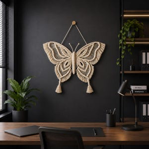 May include: A beige macrame butterfly wall hanging with detailed woven patterns. The butterfly is suspended from a wooden peg on a dark grey wall. The wings have a detailed pattern, and the piece has a bohemian aesthetic.