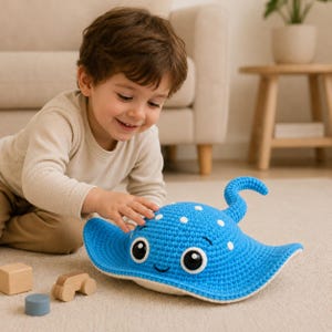May include: A blue crocheted stingray toy with white polka dots and a smiling face. The toy has a curved tail and is lying on a beige carpet. Wooden toy blocks are in the foreground.