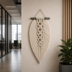 May include: A cream-colored macrame wall hanging in a leaf shape. The design features a central pattern of knotted rope with tan accents, suspended from a metal rod and a gold-colored cord. The wall hanging is displayed against a neutral-toned wall.