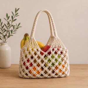 May include: A beige macrame tote bag filled with fresh fruit, including bananas, apples, oranges, and grapes. The bag has a woven design and braided handles. The bag is sitting on a wooden surface.