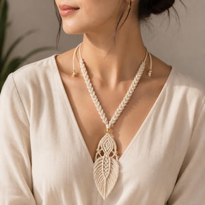 May include: Cream-coloured macrame necklace with a leaf-shaped pendant. The necklace features a braided design on the upper portion and a detailed, open-weave pattern on the pendant. Gold-coloured beads accent the design.