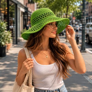 Peut inclure: Un chapeau de soleil vert vif, à tissage ouvert, avec un large bord. Le chapeau est en crochet. La personne porte un débardeur blanc et porte un sac fourre-tout beige. L'arrière-plan est une rue de la ville.