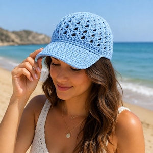 Puede incluir: Gorra de béisbol de ganchillo azul claro con visera. La gorra tiene un diseño de tejido abierto en la corona. La persona de la imagen lleva la gorra en un día soleado en la playa.