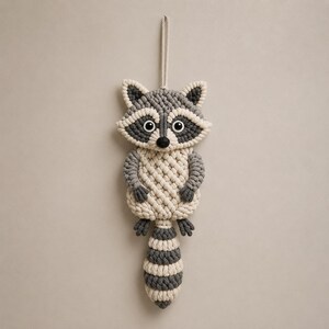May include: A handmade macrame raccoon wall hanging. The raccoon features a gray and cream color scheme with black eyes and a striped tail. It is suspended by a cream-colored cord, perfect for nursery decor or a child's room.