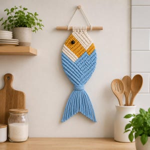 Könnte beinhalten: Makramee-Fisch-Wandbehang in Blau, Gelb und Creme. Der Fisch hat ein strukturiertes Design, ein schwarzes Auge und einen blauen Schwanz. Er hängt an einer Holzstange mit einer cremefarbenen Kordel. Der Fisch ist etwa 38 cm lang.