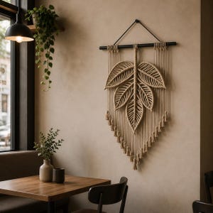 May include: A beige macrame leaf wall hanging. The leaf design is suspended from a black rod, with long, vertical strands and tassels at the bottom. The wall hanging is displayed on a beige wall, creating a natural aesthetic.