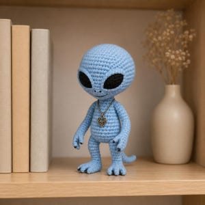 Op de afbeelding: Een lichtblauwe gehaakte alienfiguur met grote zwarte ogen en een kleine zilveren hartvormige hanger. De alien staat op een houten plank naast een vaas met gedroogde bloemen en een stapel boeken. De figuur heeft een glimlachende uitdrukking.
