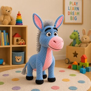 May include: A blue and pink crocheted donkey toy with large ears and a gray mane stands on a dotted rug. The toy has black hooves and a pink nose. A wooden shelf with toys and a framed sign that says "PLAY LEARN DREAM BIG" are in the background.