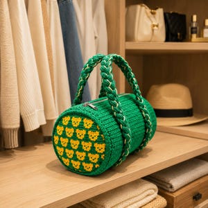 May include: A green crochet barrel bag with yellow bear face pattern on the ends. The bag has braided handles and a zipper closure. The bag is displayed on a wooden shelf in a closet.