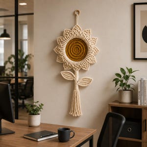 May include: A macrame sunflower wall hanging with a cream-colored petal design and a golden-yellow center. The sunflower has a stem with leaves and a tassel. It is hanging on a beige wall.