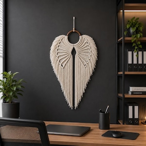 May include: A beige macrame wall hanging in the shape of angel wings. The wings are made of woven cotton rope and hang from a wooden ring. The wall decor is displayed against a dark gray wall.