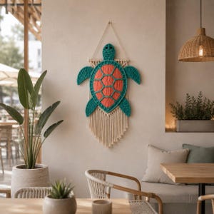 May include: A handcrafted wall hanging featuring a turtle design. The turtle has a teal outline and head, with an orange shell. The macrame art piece is suspended from a natural-colored cord and has a fringe at the bottom. It is displayed on a neutral-colored wall.