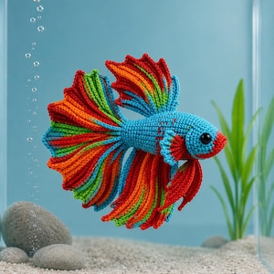 May include: A colourful crocheted Betta fish displayed in a glass aquarium. The fish has a blue body and rainbow-coloured fins. The aquarium features a sandy base, rocks, and artificial plants, with bubbles rising in the water.