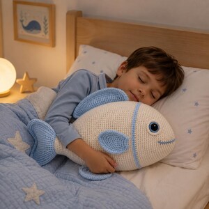Op de afbeelding: Een crème en blauw gehaakt visvormig kussen. Het kussen heeft blauwe vinnen en een lachend gezicht. Het kussen wordt vastgehouden door een persoon in een blauw shirt, liggend op een bed met een blauw dekbed en een kussen met blauwe sterren.