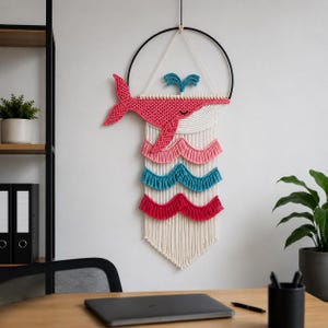 May include: A handcrafted wall hanging featuring a pink crocheted whale with a blue spout. Below the whale are layers of pink, teal, and red fringe. The wall decor is suspended from a black metal ring.