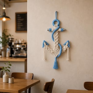 Può includere: Un'ancora in macramè da parete a tema nautico. L'ancora è realizzata con corda bianca e blu, con un design a nodi dettagliato. Presenta un nappa blu sul fondo ed è montata su una parete neutra, creando un'estetica costiera.