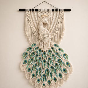 May include: Cream-coloured macrame peacock wall hanging with teal, blue, and green accents. The peacock has a detailed tail and is suspended from a black rod.