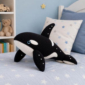 May include: A crocheted orca whale toy in black and white, with white spots and blue eyes. The soft toy is on a light blue bedspread with a star pattern. The orca has a white underside and a black dorsal fin. A children's bedroom setting.