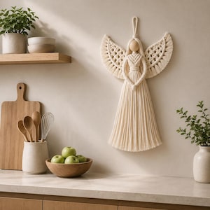 May include: Cream-colored macrame angel wall hanging with outstretched wings. The angel features a wooden head and a flowing skirt-like design. The wings have a detailed woven pattern. The angel is suspended by a loop, suitable for hanging. The background is a neutral-toned wall.