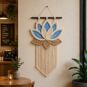 May include: A macramé wall hanging in the shape of a lotus flower. The flower has blue and beige petals, a beige center, and a fringe. It is suspended from a black rod and displayed on a neutral wall.