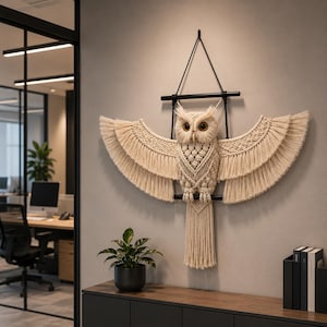 May include: A macrame owl wall hanging with outstretched wings. The owl is made of cream-colored woven cotton, with a detailed body and fringed wings. It hangs from a black metal frame, creating a decorative and artistic wall accent.