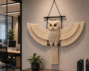 May include: A macrame owl wall hanging with outstretched wings. The owl is made of cream-colored woven cotton, with a detailed body and fringed wings. It hangs from a black metal frame, creating a decorative and artistic wall accent.